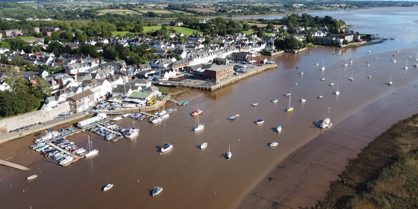 Aerial view of a waterfront town on a wide estuary, with boats moored offshore and green countryside beyond.
