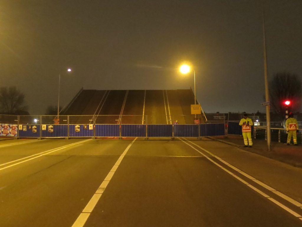 A road closed at night behind temporary fencing and barriers, with a raised lifting bridge beyond and workers in high-visibility clothing.