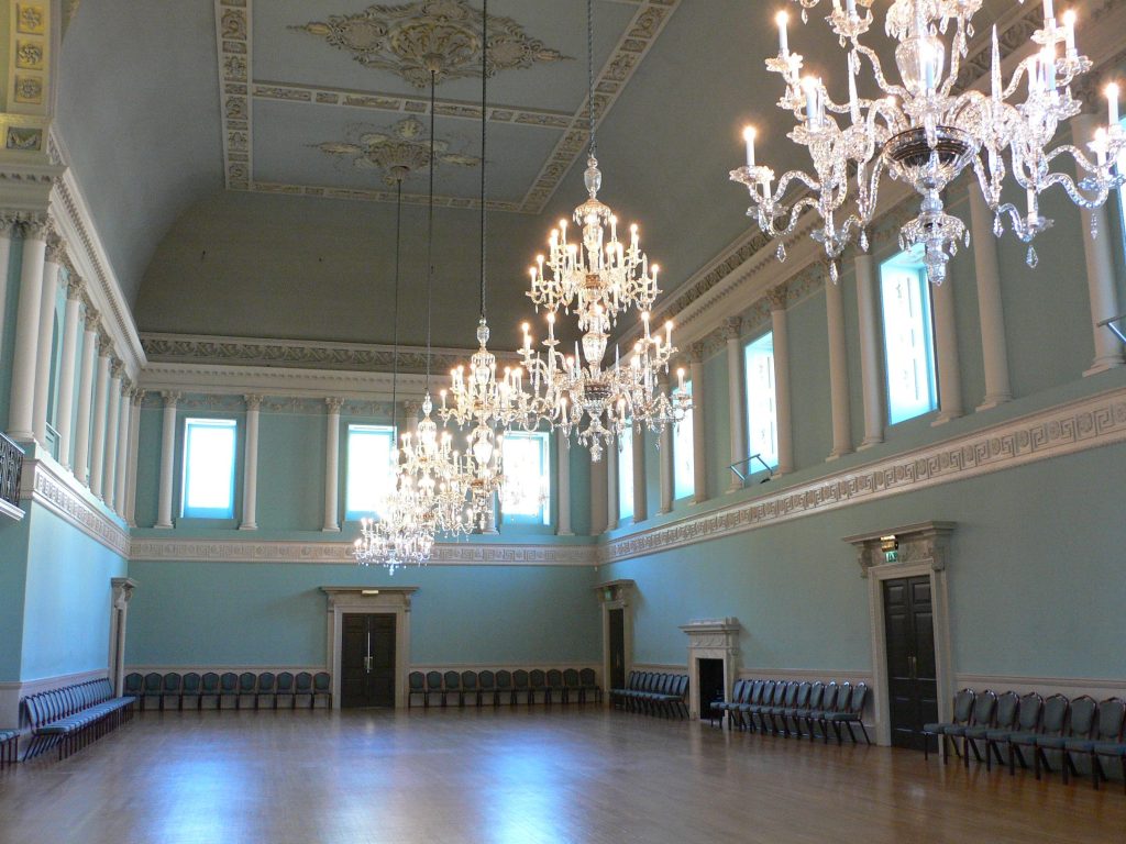 An elegant empty hall with crystal chandeliers, tall windows and chairs set along the walls, ready for a ceremony.