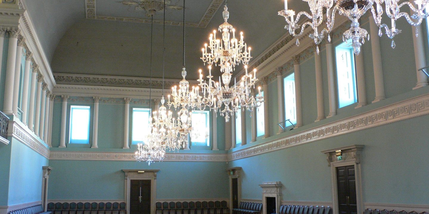 An elegant empty hall with crystal chandeliers, tall windows and chairs set along the walls, ready for a ceremony.