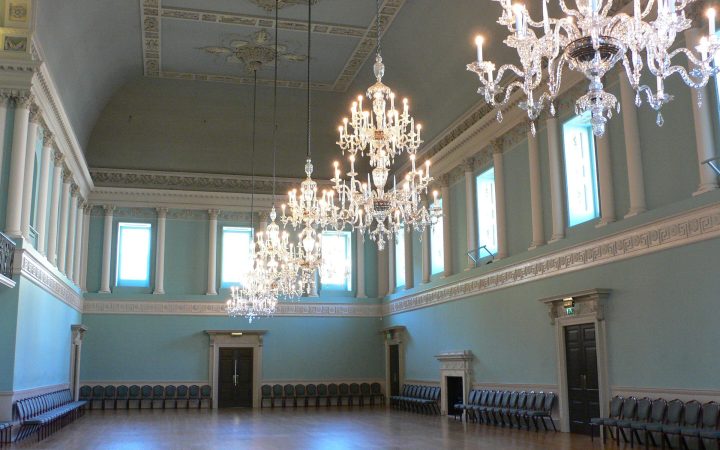 An elegant empty hall with crystal chandeliers, tall windows and chairs set along the walls, ready for a ceremony.