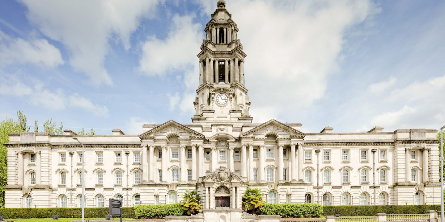 A grand white stone civic building with a central clock tower, columns and formal lawns in front.