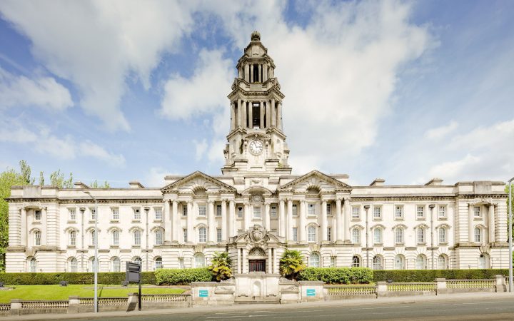 A grand white stone civic building with a central clock tower, columns and formal lawns in front.