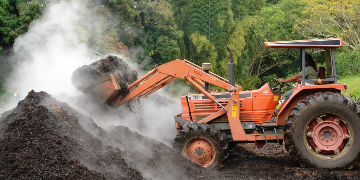 A tractor with a front loader turning a large steaming heap of compost.