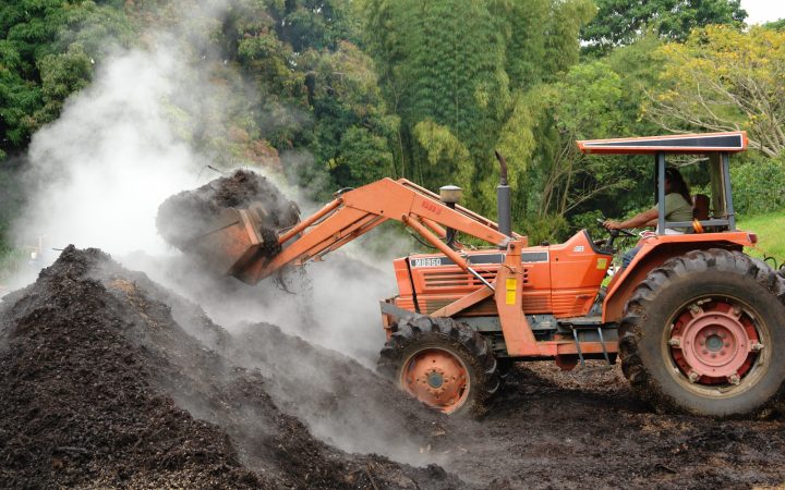 A tractor with a front loader turning a large steaming heap of compost.