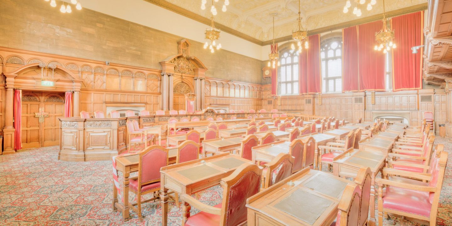 An empty council chamber with rows of wooden desks and red leather chairs facing a panelled dais, lit by brass chandeliers.