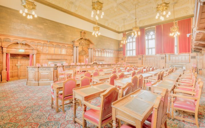 An empty council chamber with rows of wooden desks and red leather chairs facing a panelled dais, lit by brass chandeliers.