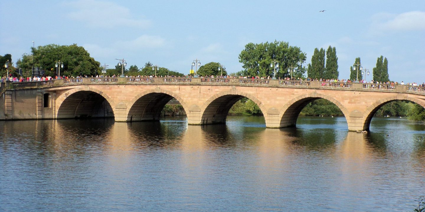 An arched stone road bridge spanning a calm river on a bright, sunny day.