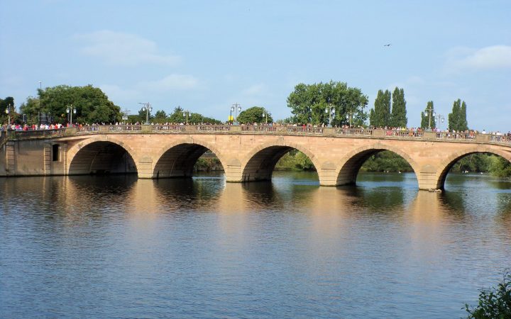 An arched stone road bridge spanning a calm river on a bright, sunny day.
