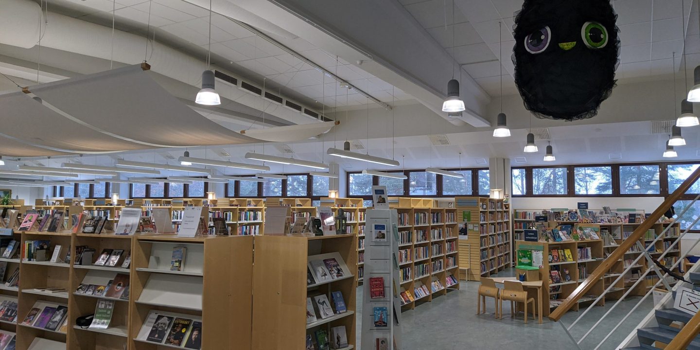 A bright, open-plan public library with rows of wooden bookshelves, display stands and windows along the far wall.