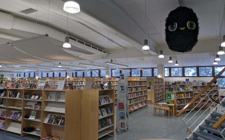 A bright, open-plan public library with rows of wooden bookshelves, display stands and windows along the far wall.
