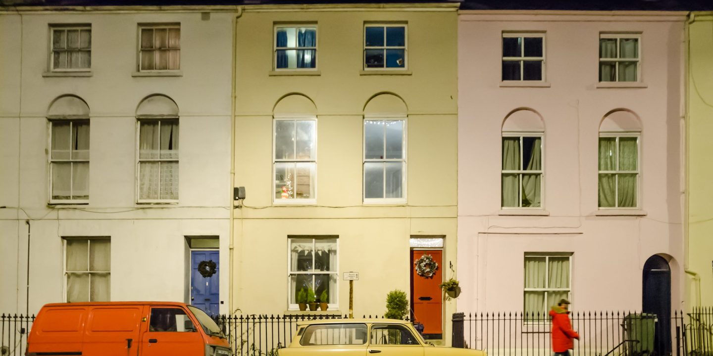 A row of pale painted terraced houses with older cars parked along the kerb.