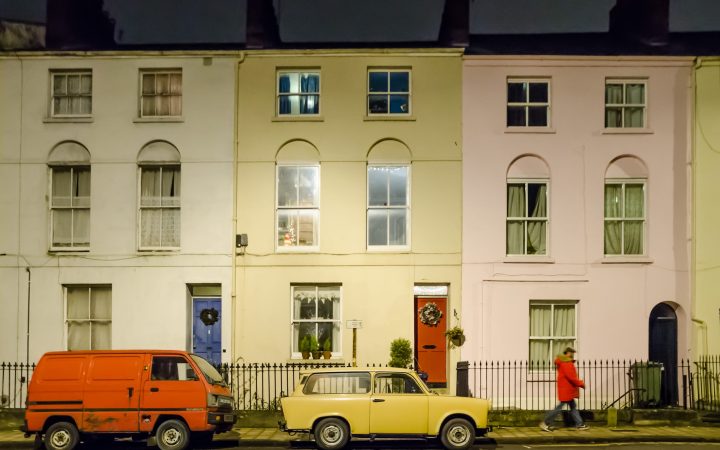 A row of pale painted terraced houses with older cars parked along the kerb.