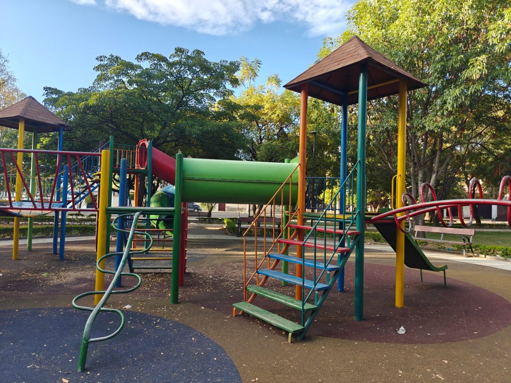 A colourful playground with climbing towers, slides and ladders on soft safety surfacing, surrounded by trees.