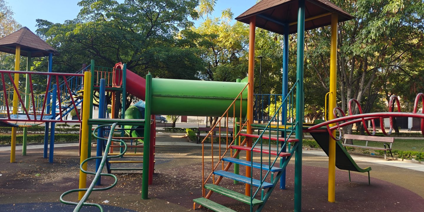 A colourful playground with climbing towers, slides and ladders on soft safety surfacing, surrounded by trees.