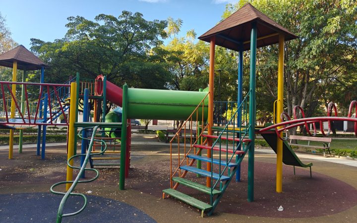A colourful playground with climbing towers, slides and ladders on soft safety surfacing, surrounded by trees.