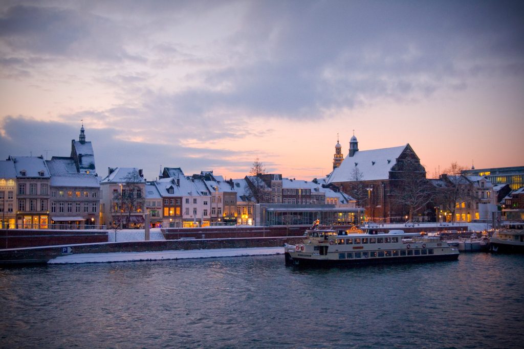 A river running through a city at dusk, with a moored boat and lit buildings along the waterfront under snow-covered roofs.
