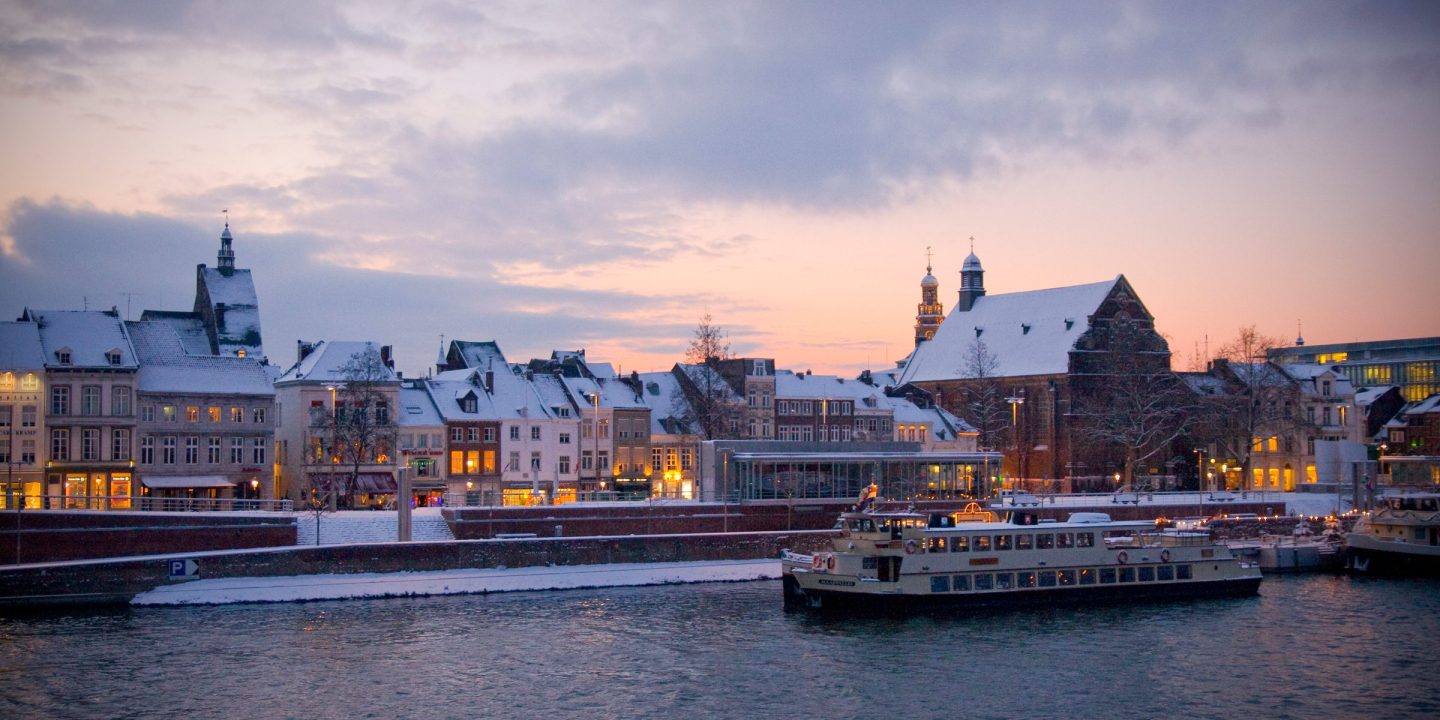 A river running through a city at dusk, with a moored boat and lit buildings along the waterfront under snow-covered roofs.