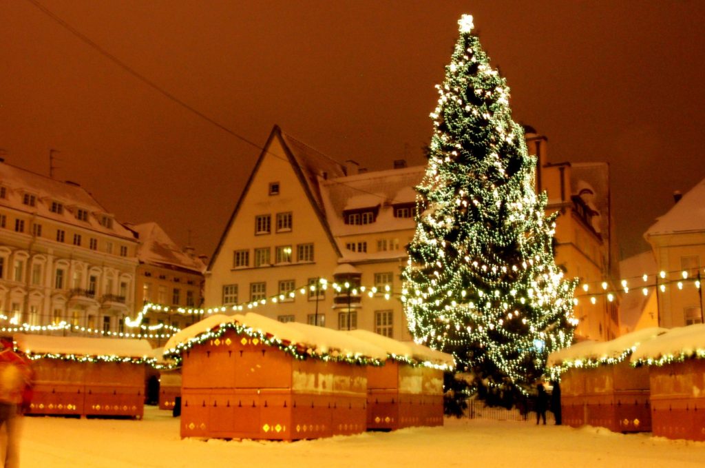 Christmas market stalls dusted with snow and strung with lights around a tall illuminated Christmas tree at night.