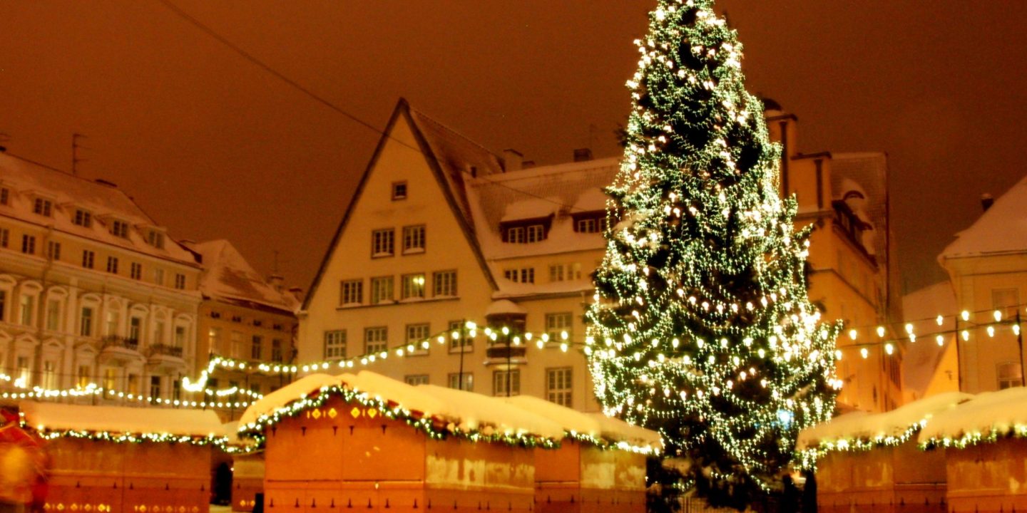 Christmas market stalls dusted with snow and strung with lights around a tall illuminated Christmas tree at night.