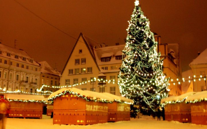 Christmas market stalls dusted with snow and strung with lights around a tall illuminated Christmas tree at night.