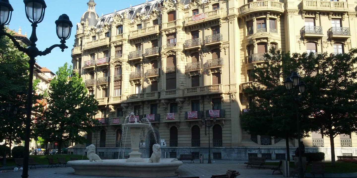 A paved city square with a stone fountain, benches and old-style lamp posts in front of an ornate building.