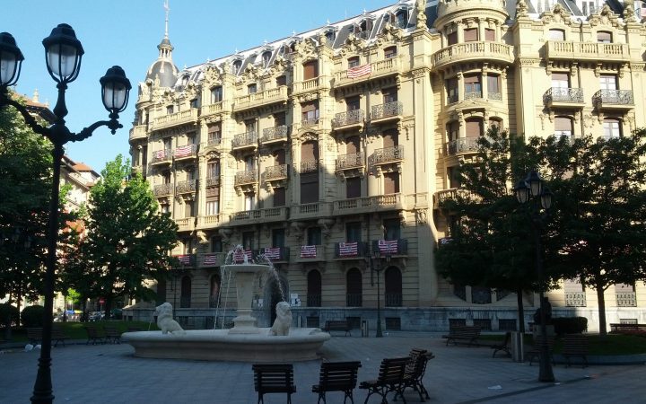 A paved city square with a stone fountain, benches and old-style lamp posts in front of an ornate building.