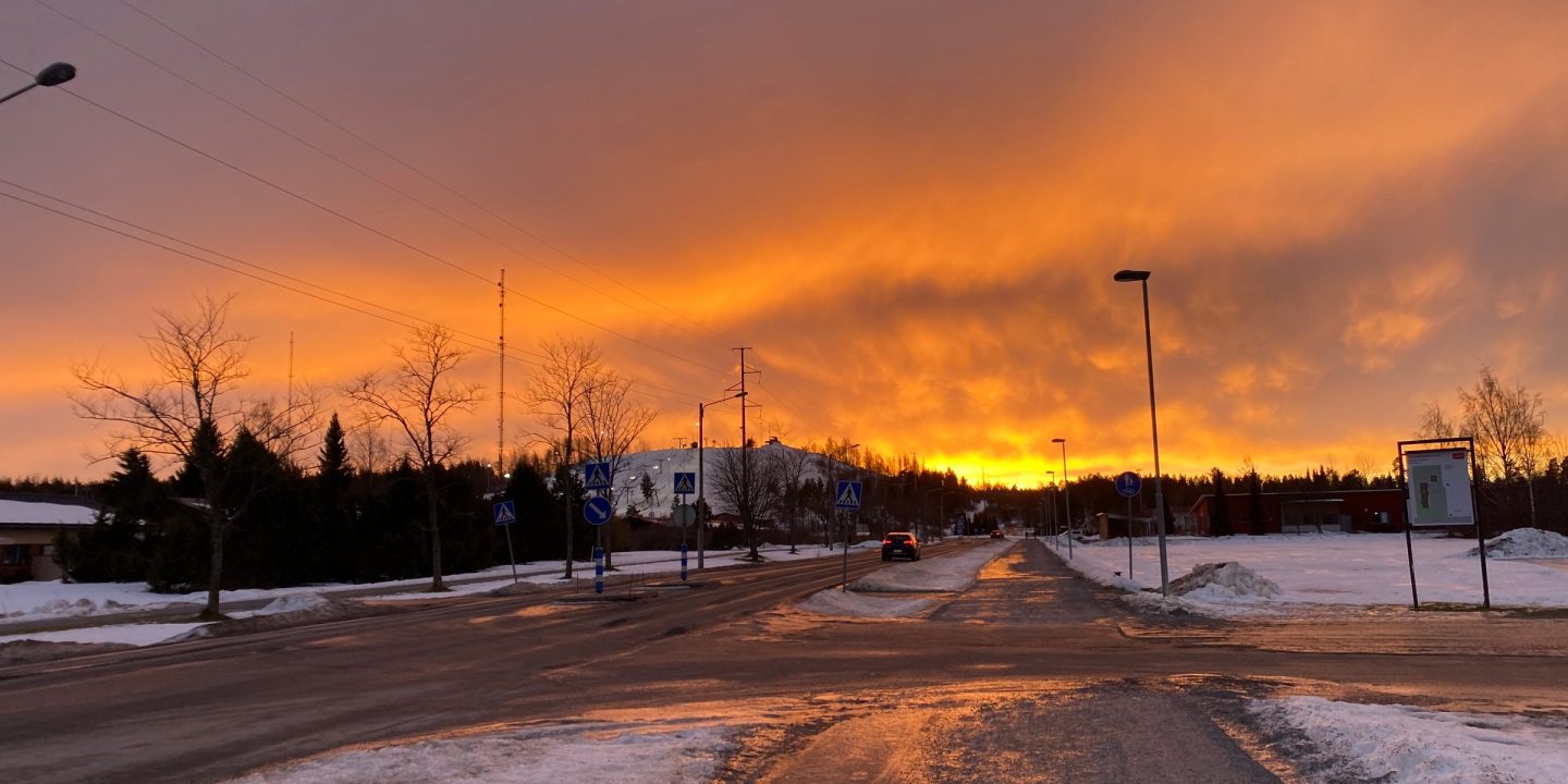 A snowy street at sunset, with a fiery orange sky behind bare trees and street lights.