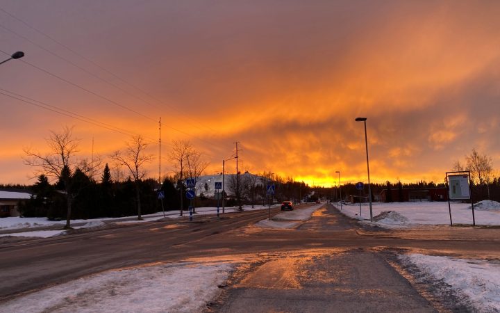 A snowy street at sunset, with a fiery orange sky behind bare trees and street lights.