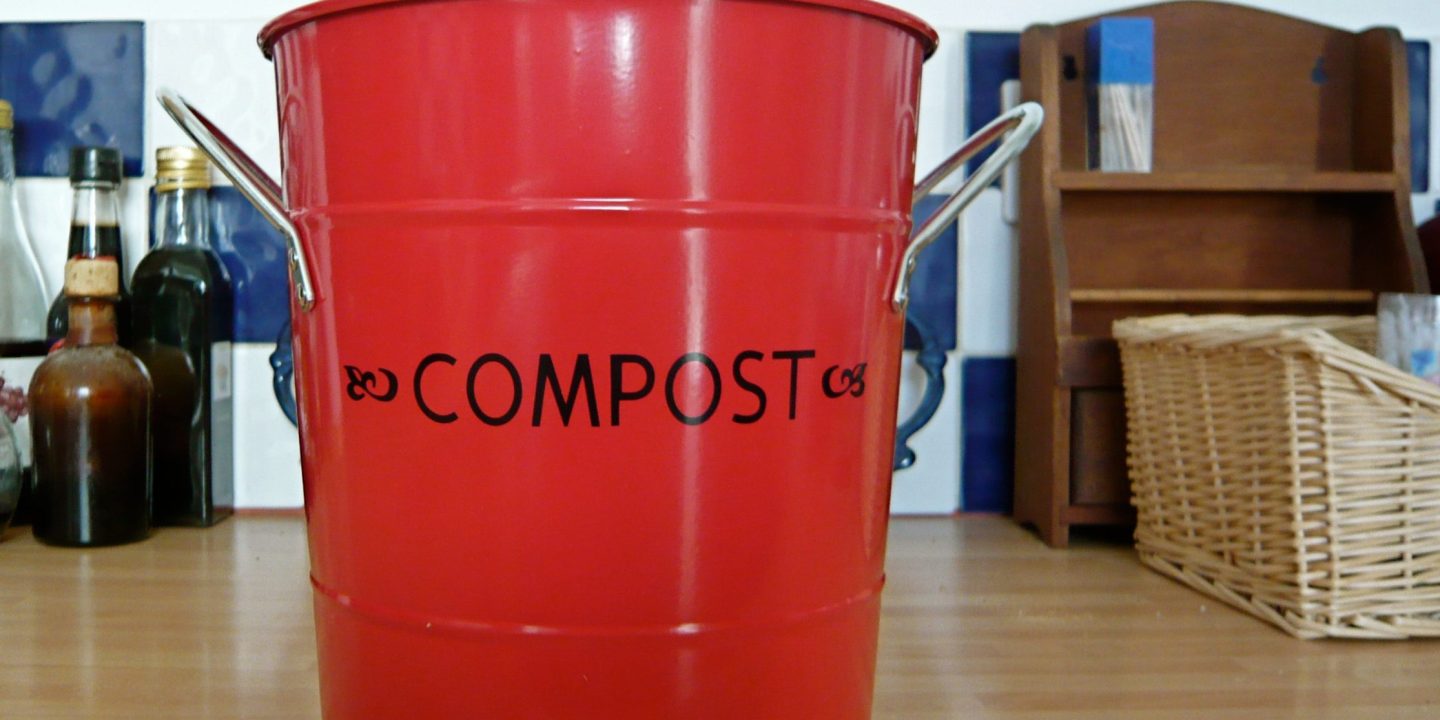 A red metal kitchen caddy labelled 'Compost' standing on a kitchen worktop.