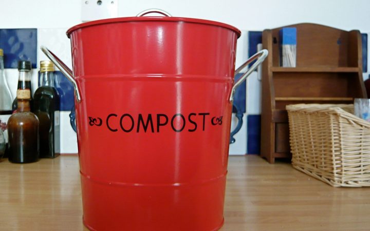 A red metal kitchen caddy labelled 'Compost' standing on a kitchen worktop.