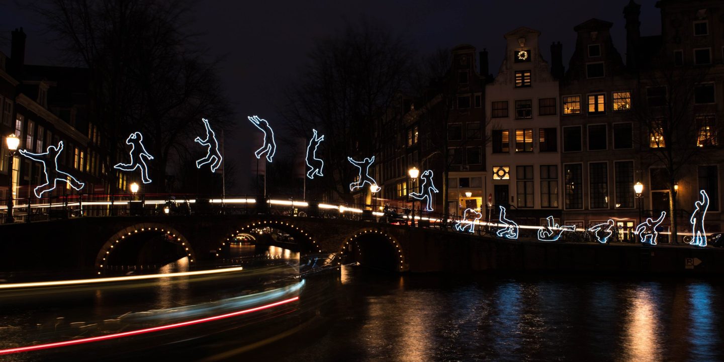 A light art installation at night: glowing outlines of leaping figures above a bridge, with light trails from a passing boat reflected in the water.