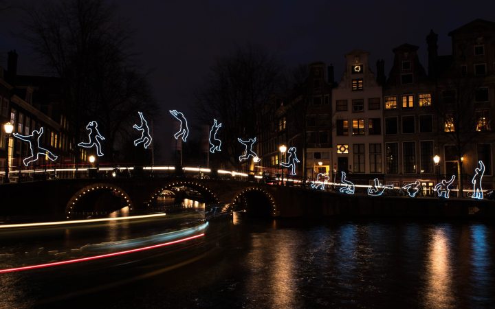 A light art installation at night: glowing outlines of leaping figures above a bridge, with light trails from a passing boat reflected in the water.